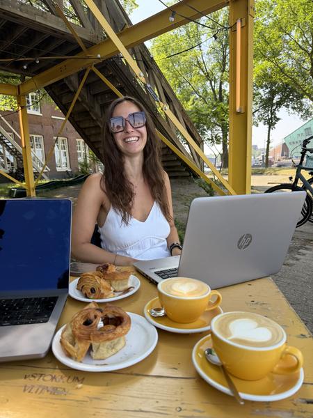 A person is happily working outdoors at a yellow wooden table, enjoying coffee and pastries